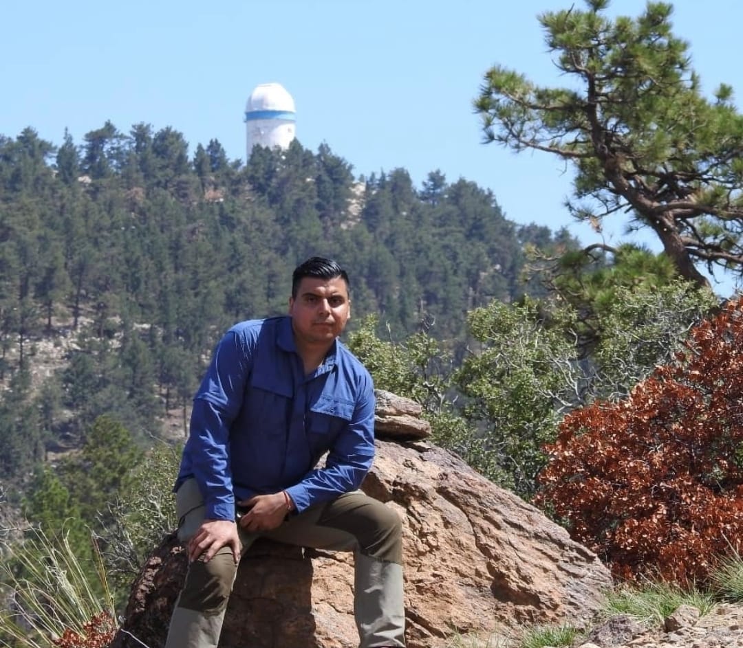 Miguel Leon, sitting on a rock with a scenic background of trees and an observatory tower.