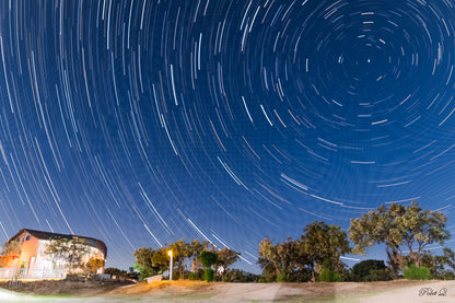 Star trail effect over a rural landscape with trees and a building.