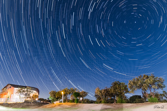 Star trail effect over a rural landscape with trees and a building.